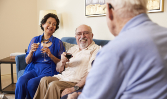 A couple sits on a couch with drinks, talking with a friend in the foreground.