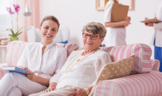 A senior woman and younger woman are in assisted living, smiling and sitting on a couch, memory care Montrose, CO