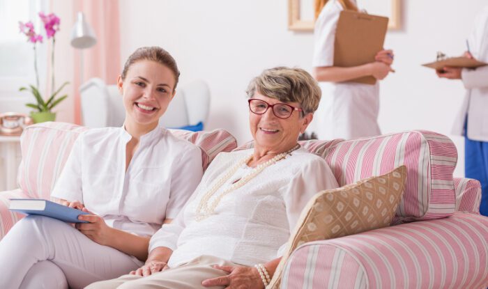 A senior woman and younger woman are in assisted living, smiling and sitting on a couch, memory care Montrose, CO
